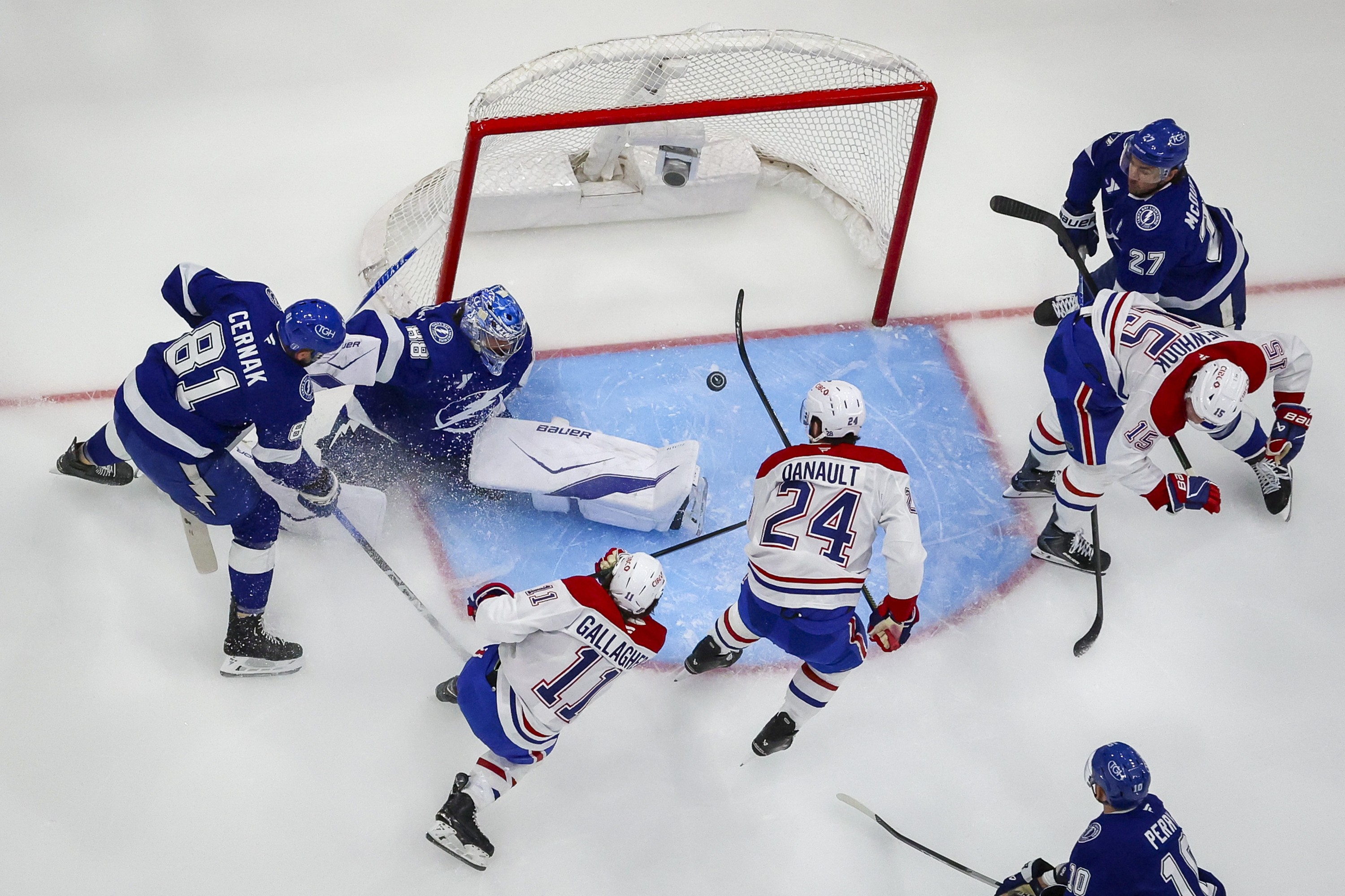 An overhead photo shows the puck mid-air in front of a mostly empty net as Brendan Gallagher follows through on a shot and several other players are around the crease.