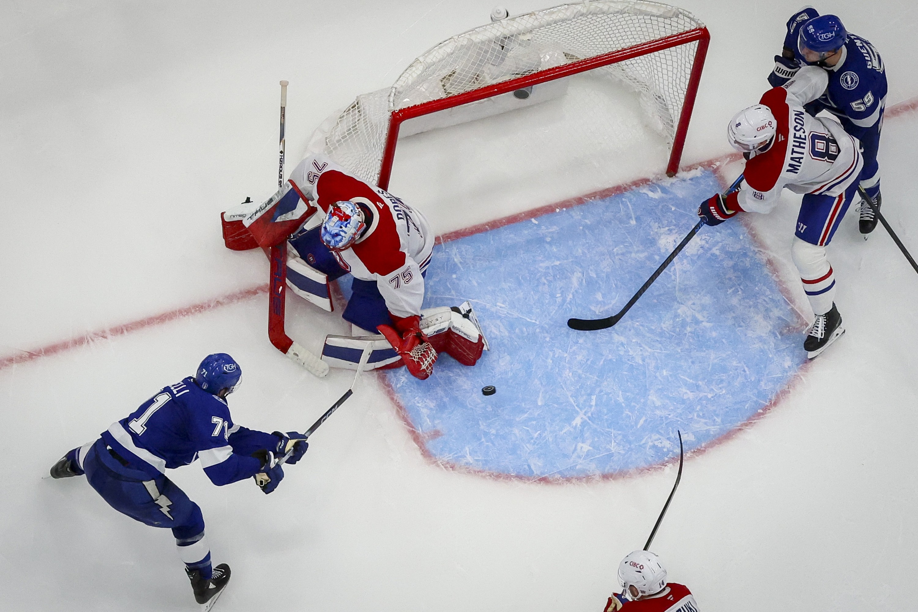 The puck is mid-air in the crease next to Canadiens goaltender Jakub Dobes as Lightning's Anthony Cirelli follows through on a shot and other players gather around the crease.