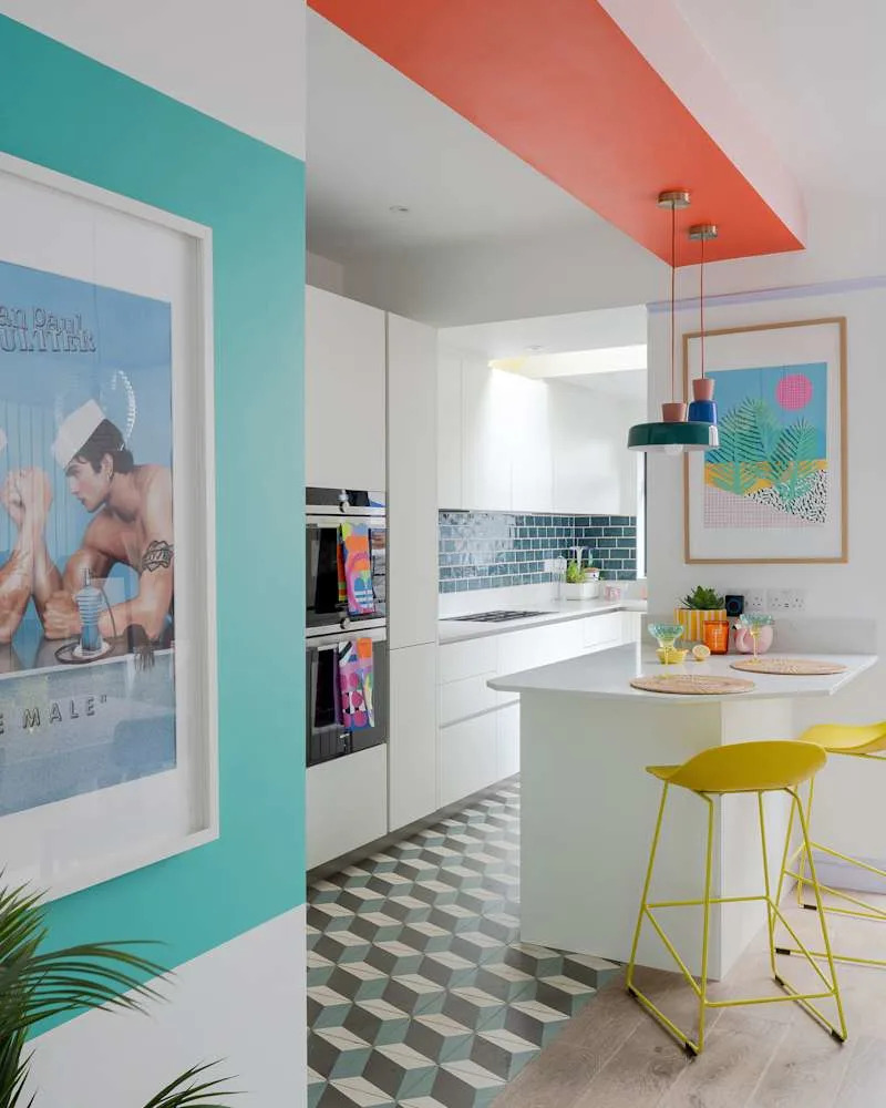 Bright kitchen with teal and coral accents, geometric floor tiles, white cabinets, and colorful bar stools.