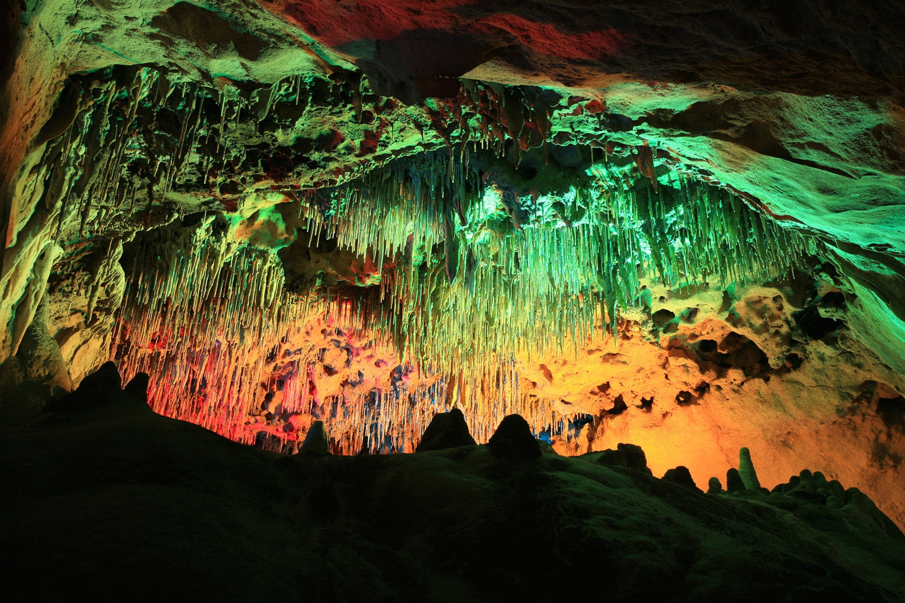 Cave interior with stalactites hanging from ceiling is lit with colorful green, red, and orange lights