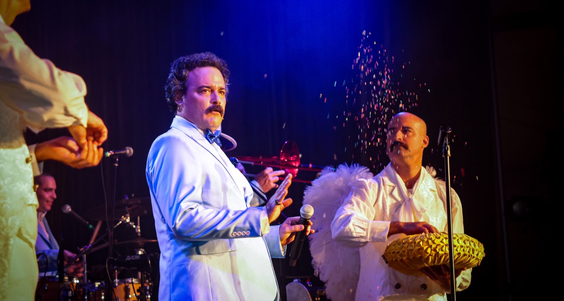 Photo of a an actor onstage paying comedian Alvarez Guedes. Next to him is a man dressed as an angel throwing glitter in the air