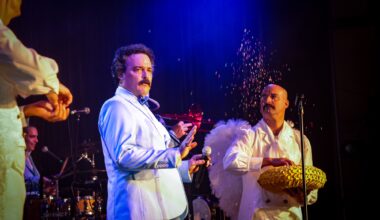 Photo of a an actor onstage paying comedian Alvarez Guedes. Next to him is a man dressed as an angel throwing glitter in the air