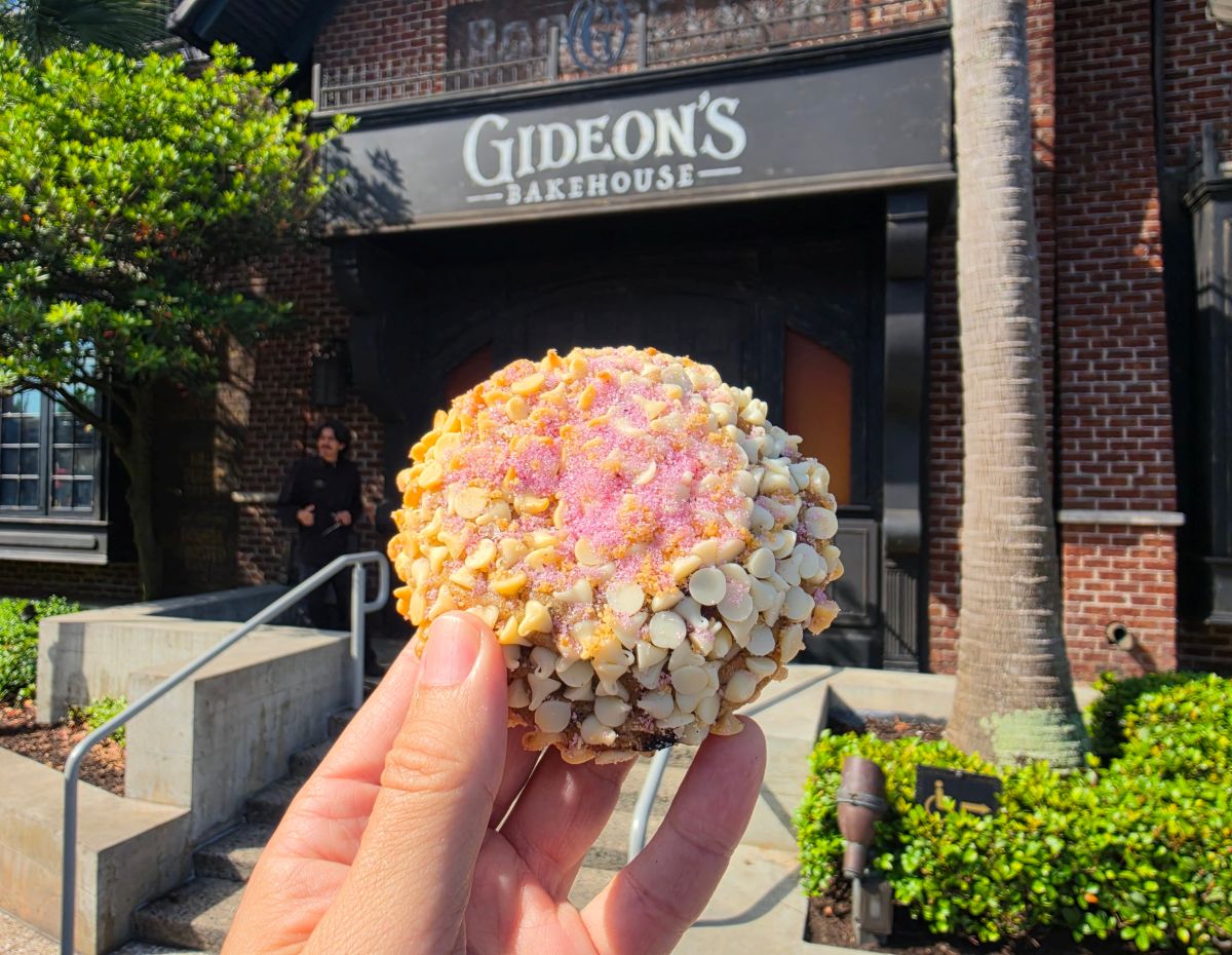 A close-up of a large, thick cookie covered in yogurt chips and pink sugar sparkles from Gideon's Bakehouse at Disney Springs, held up in front of the bakery's storefront.
