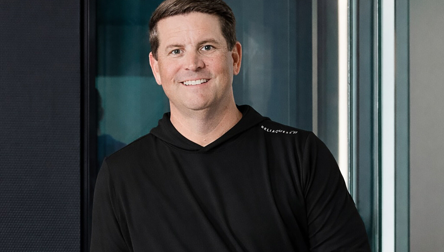 Man in black hoodie posed in front of glass wall.