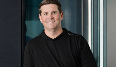 Man in black hoodie posed in front of glass wall.