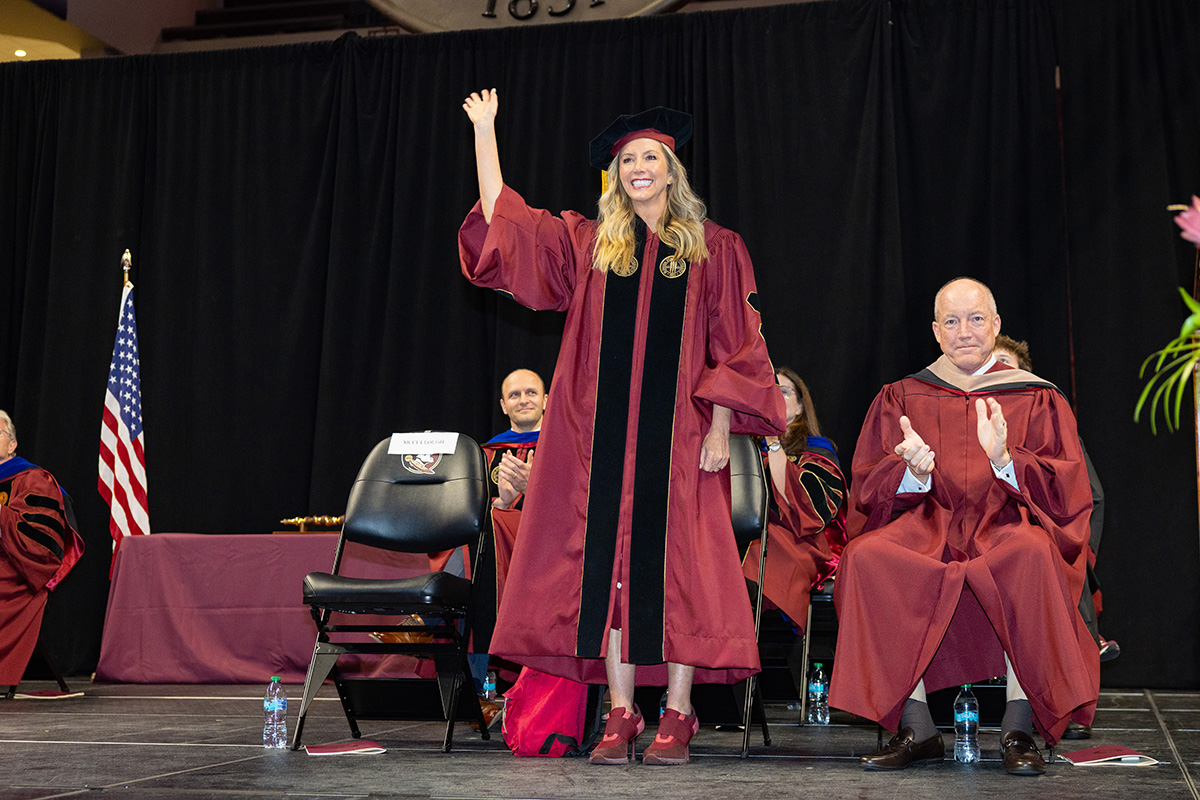 Sara Blakely, in a garnet graduation robe, waves to the crowd on stage during a commencement ceremony.