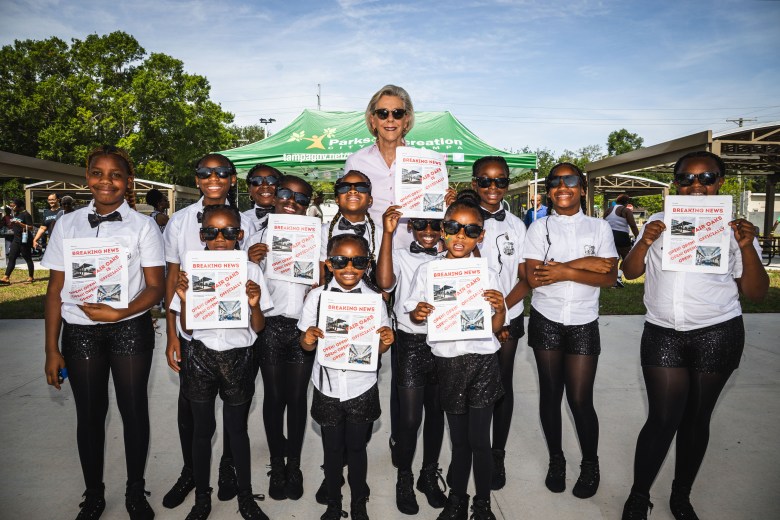 A bright, outdoor daytime photograph of Tampa Mayor Jane Castor posing with a group of about ten smiling children at the Fair Oaks Recreation Complex. Mayor Castor stands in the back center, wearing sunglasses and a white top. The children, arranged in two rows in the foreground, are uniformly dressed in white button-down shirts, black bow ties, black pants, and dark sunglasses. Several of the children are proudly holding up white flyers or certificates. The group is standing on a paved surface under a park pavilion structure, with a green event tent, mature trees, and a clear blue sky visible in the background.