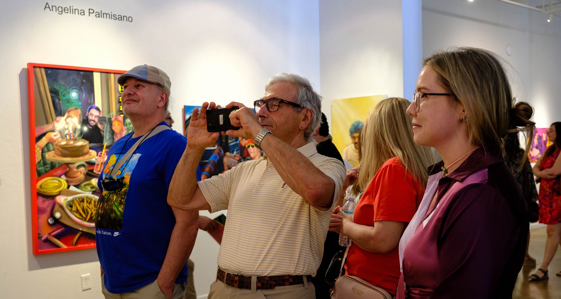 People attending an art gallery exhibition, with several observing and photographing colorful paintings on white walls.