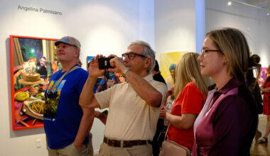 People attending an art gallery exhibition, with several observing and photographing colorful paintings on white walls.