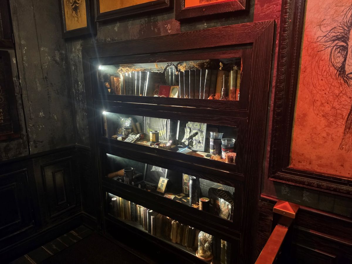A tall, dark wood display cabinet, lit from within, at Gideon's Bakehouse, showcasing old books and vintage-style merchandise beneath a gallery wall of gothic art.