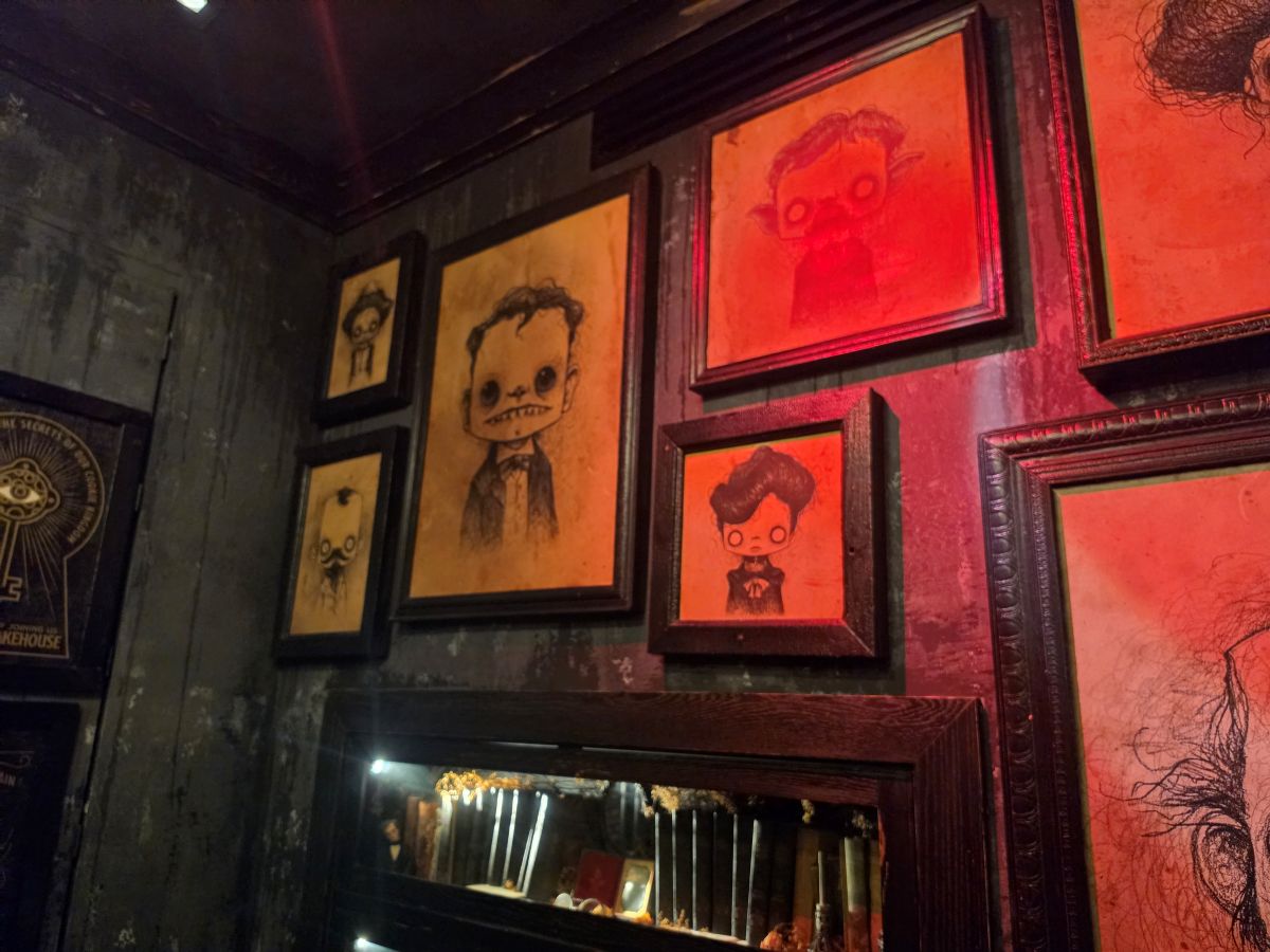 Moody interior gallery wall at Gideon's Bakehouse featuring framed, spooky character portraits washed in eerie red and sepia lighting above a display shelf.