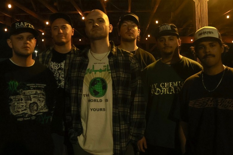 A low-light, nighttime group photograph of the six members of the band Holy Dose standing together outdoors. The band is arranged in a casual, staggered line facing the camera against a very dark background illuminated only by softly blurred string lights overhead. The members are dressed in casual streetwear, with most wearing baseball caps and dark graphic t-shirts. In the center foreground, one member with a shaved head is wearing an open plaid flannel shirt layered over a light-colored t-shirt with a globe graphic and the text 'THE WORLD IS YOURS'. The overall lighting is dim and heavily shadowed, creating a raw and gritty atmosphere.