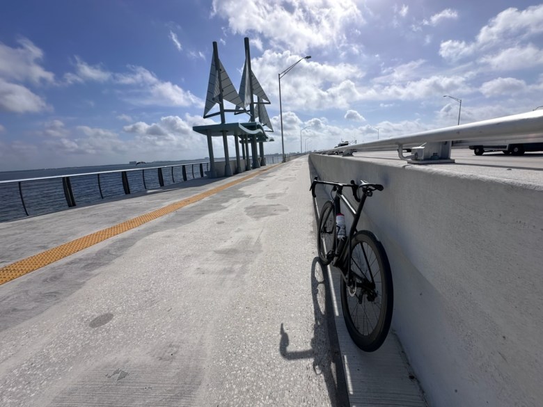 A bicycle leans against the wall of the Howard Franklin Bridge Trail with a sailboat-shaped shade and sunny, cloudy skies in the background.