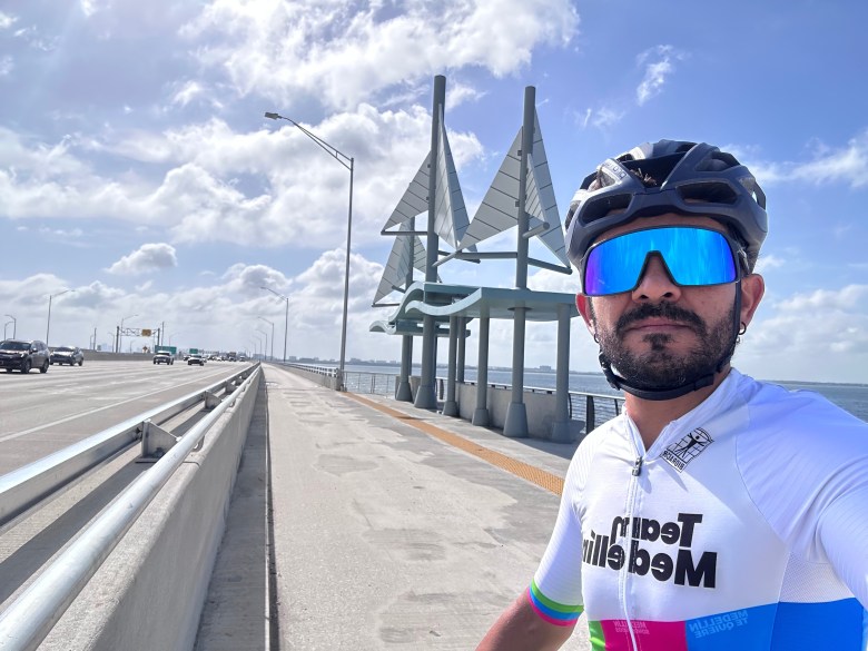 Man in reflective sunglasses and a cycling jersey takes a selfie in front of the sailboat shaped shade stop on the Howard Franklin Bridge trail