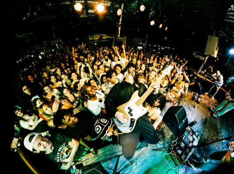 "A high-energy, fisheye-lens live concert photograph taken from the stage, looking out into a packed, enthusiastic crowd at Crowbar. In the center foreground, a musician is dynamically playing a white electric guitar or bass. The dense audience is pressed right up against the stage, with dozens of attendees cheering, smiling, and raising their hands in the air. In the bottom left foreground, an attendee wearing a backward white baseball cap looks up toward the performer. The dark, intimate venue is illuminated by bright overhead stage lights, capturing the intense, lively atmosphere of a punk or indie rock show.