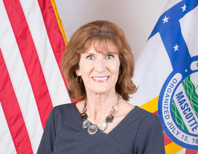 A formal, close-up studio portrait of Jean Duncan, the Administrator of Infrastructure & Mobility for the City of Tampa. They have shoulder-length reddish-brown hair, hoop earrings, and are smiling warmly at the camera. They are wearing a dark, short-sleeved v-neck top accompanied by a distinct statement necklace made of large, flat metallic and stone-like discs. In the background, they are flanked by the United States flag on the left and a municipal flag featuring a city seal on the right against a plain, light-colored wall.