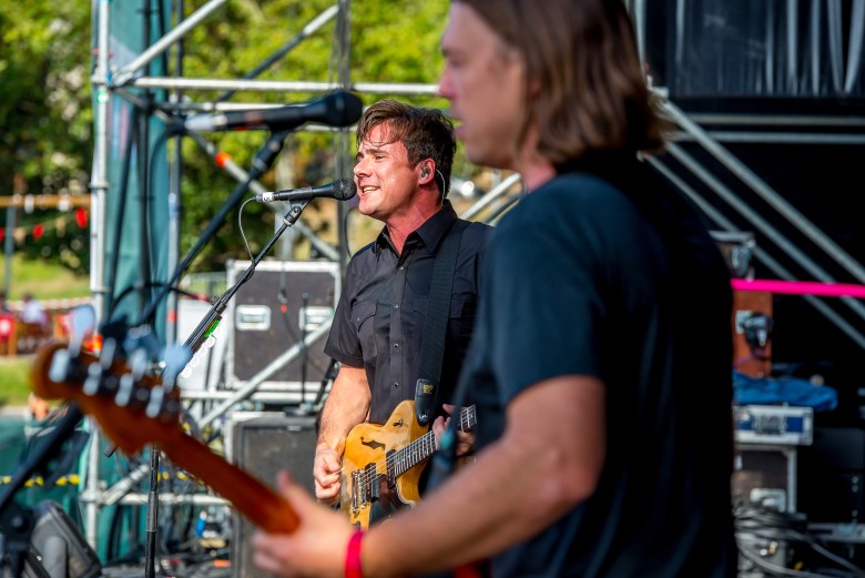 A daytime live concert photograph of Jim Adkins from the rock band Jimmy Eat World performing on an outdoor stage. Adkins is in sharp focus in the background, singing passionately into a black microphone while playing a light wood-colored electric guitar. They have short brown hair and are wearing a black short-sleeve button-down shirt. In the blurred foreground to the right, the back and profile of another band member playing a reddish-brown bass guitar is visible. The scene is brightly lit by natural sunlight, with metal stage scaffolding and green foliage visible in the background.