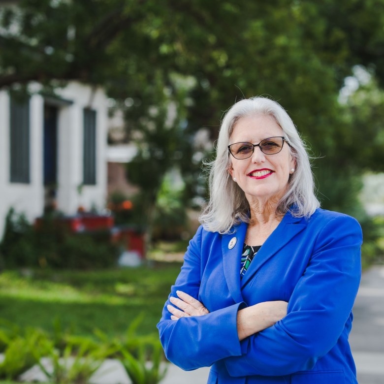 A daytime outdoor portrait of former Hillsborough County Commissioner Kimberly Overman. They are a white individual with shoulder-length silver hair, wearing dark-framed glasses and bright red lipstick. Overman is smiling warmly at the camera with their arms crossed, wearing a bright royal blue blazer over a dark patterned top. The background is softly blurred with a shallow depth of field, revealing a sunlit residential area with green trees, a sidewalk, and a white building.