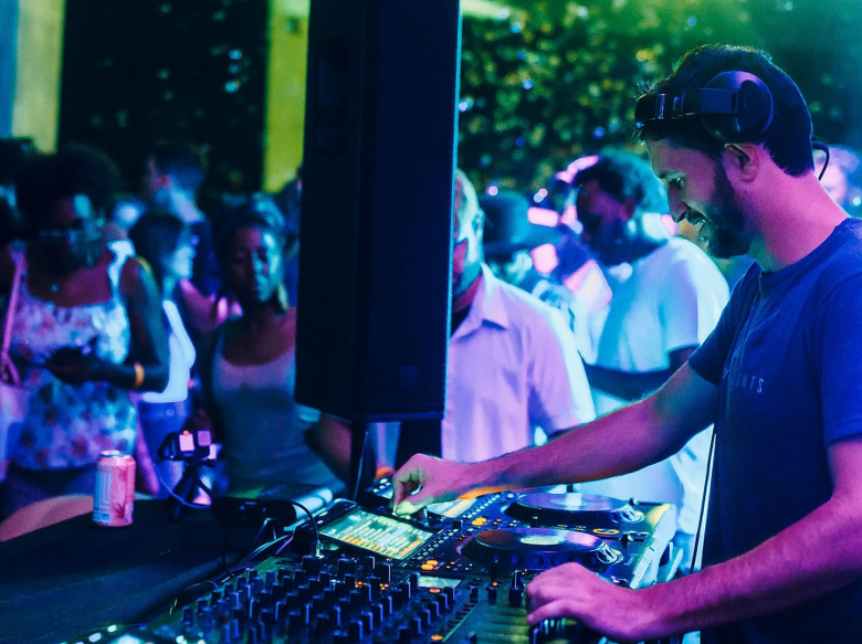 A vibrant, nighttime live performance photograph of electronic musician Taetro DJing at the St. Pete Pier. In the right foreground, Taetro has short dark hair and a beard, wearing a blue t-shirt with over-ear headphones resting on their head. They are looking down intently while using both hands to adjust the knobs and sliders on a large, illuminated DJ mixer controller. A tall black speaker stands vertically in the center of the frame, separating the DJ booth from the audience. In the softly blurred background to the left, a crowd of partygoers is visible mingling and dancing. The entire scene is heavily illuminated by bright, moody blue and cyan stage lighting.