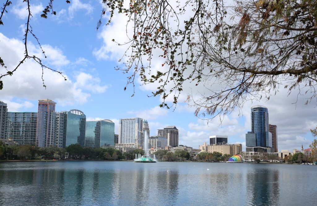 view across Lake Eola shows iconic fountain and Orlando skyline against blue sky, white clouds, and tree leaves