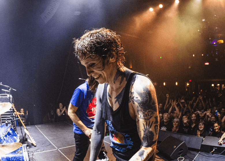 A wide live concert photograph of Laura Jane Grace & The Mississippi Medicals performing on a dimly lit stage. In the center foreground, Laura Jane Grace, featuring dark curly hair and heavily tattooed arms, smiles widely while playing a black electric guitar and wearing a black tank top. The scene is illuminated by striking white beams from above and warm orange stage lights, with a packed crowd visible in the background.