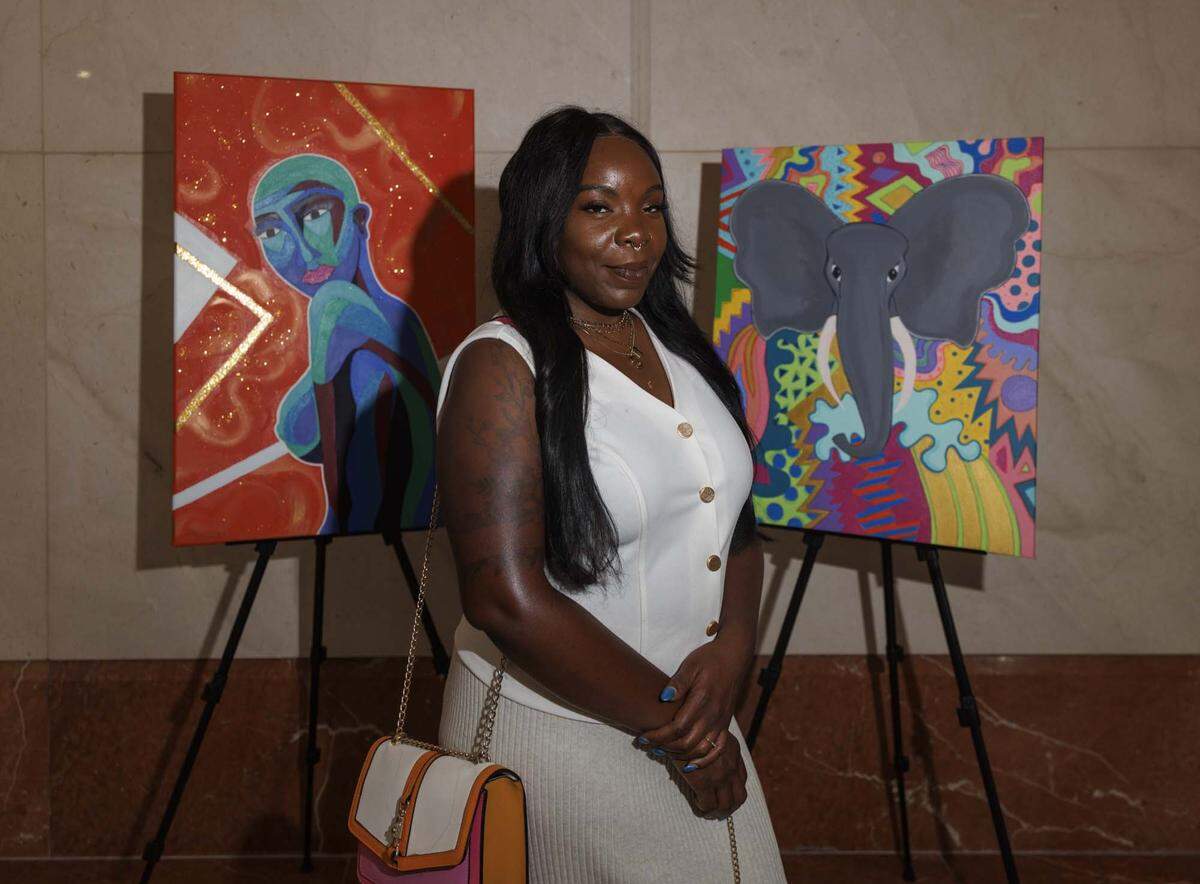Showana Louisma, poses with her art pieces, during the Camillus House art show on Wednesday, May 27, 2026, in the foyer at Amerant Bank in Coral Gables, Fla. Louisma has been at Camillus since last fall. “Honestly, on the days I want to give up, art brings me back,” said Louisma.