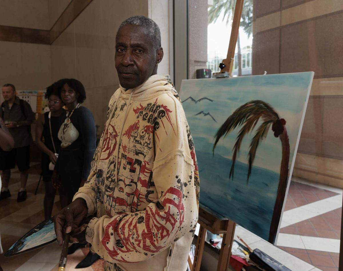 Barry Glenn, stroke survivor and artist, poses in front of his painting between working on it during the Camillus House art show on Wednesday, May 27, 2026, in the foyer at Amerant Bank in Coral Gables, Fla. Glenn says painting has helped him physically after his stroke but also mentally. “It's more of a therapy to see what can come of doing all this art,” said Glenn. 