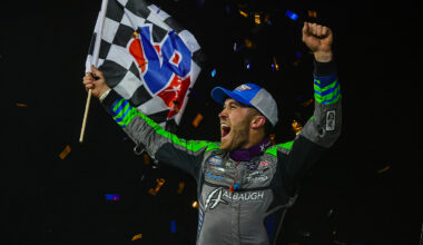Driver celebrating victory, holding a checkered flag with arms raised amid confetti against a dark background.