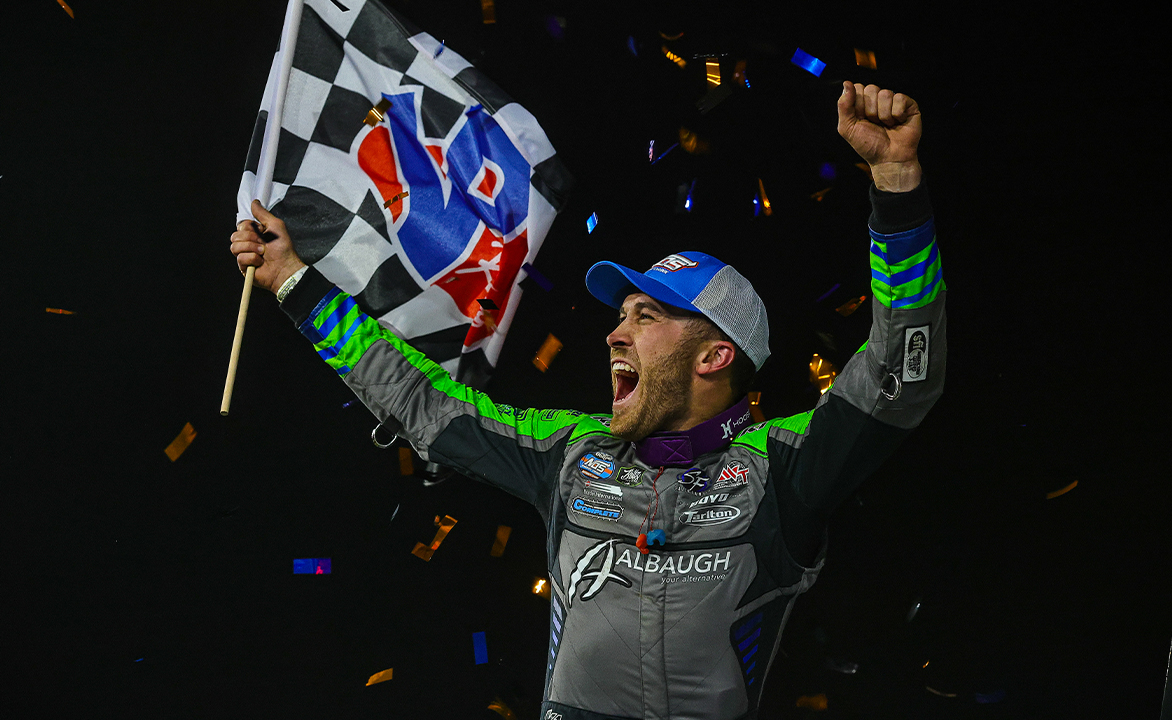 Driver celebrating victory, holding a checkered flag with arms raised amid confetti against a dark background.