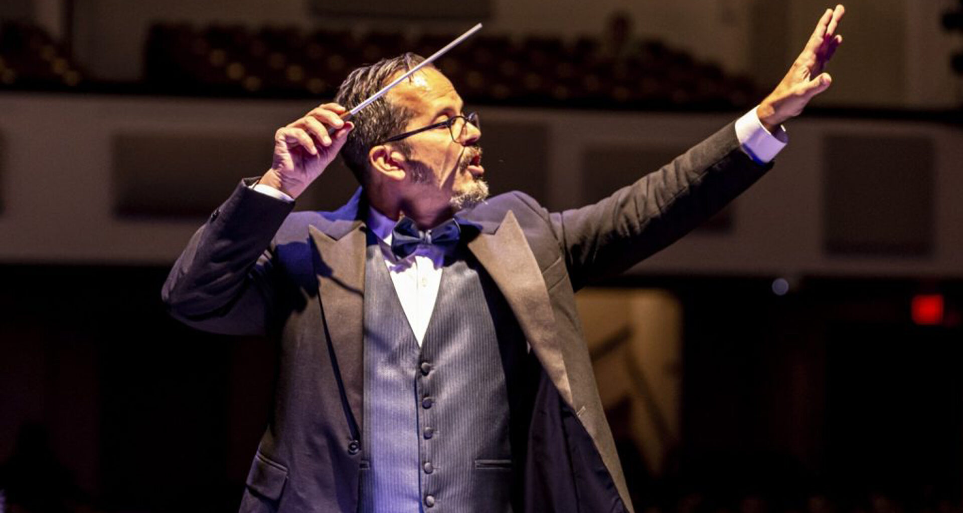 A conductor wearing a formal suit with a bow tie leads an orchestra, with a baton in his right hand and left arm raised.
