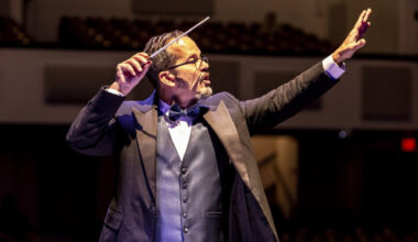 A conductor wearing a formal suit with a bow tie leads an orchestra, with a baton in his right hand and left arm raised.