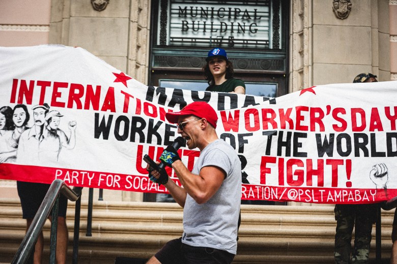A daytime photograph of an organizer, Will Kilgore from the St. Pete Tenants Union, speaking on the stone steps of the St. Petersburg City Hall during a May Day rally. Will, wearing a backward red baseball cap, glasses, and a grey t-shirt, is captured mid-speech in the foreground. Behind them, demonstrators hold a large white banner with prominent red and black text reading 'MAY DAY,' 'INTERNATIONAL WORKER'S DAY,' 'WORKERS OF THE WORLD FIGHT!' and 'PARTY FOR SOCIALISM AND LIBERATION / @PSLTAMPABAY'. The group is positioned in front of dark entryway doors with the words 'MUNICIPAL BUILDING' inscribed in stone above.
