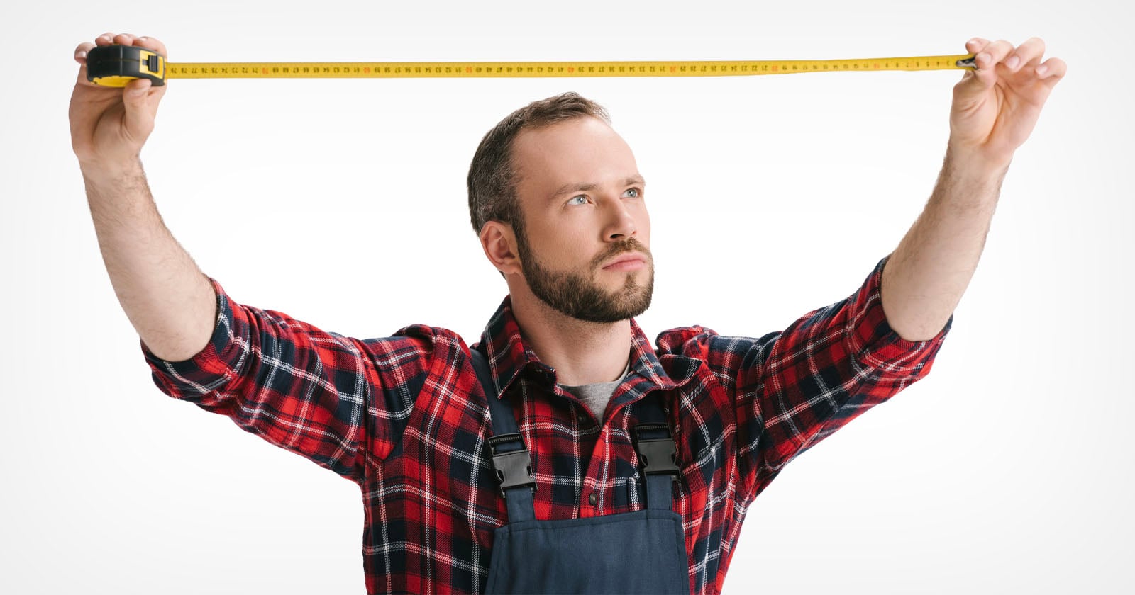 A man in a red plaid shirt and overalls holds a yellow measuring tape, stretching it out horizontally with both hands against a white background.