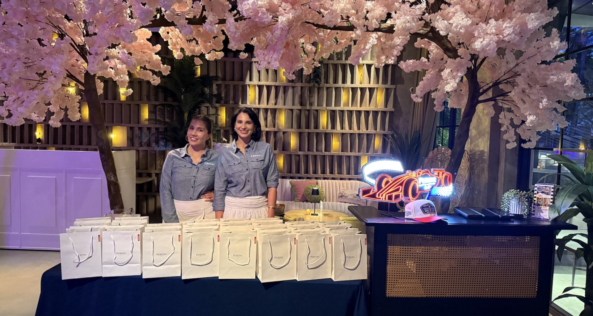 Photo of two women posing inside a bar in front of a table with white goodie bags and a tablecloth reading, "Peroni." A decorative cherry-blossom tree is seen above and behind them