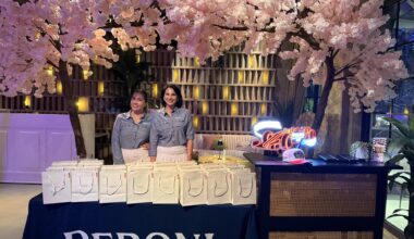 Photo of two women posing inside a bar in front of a table with white goodie bags and a tablecloth reading, "Peroni." A decorative cherry-blossom tree is seen above and behind them