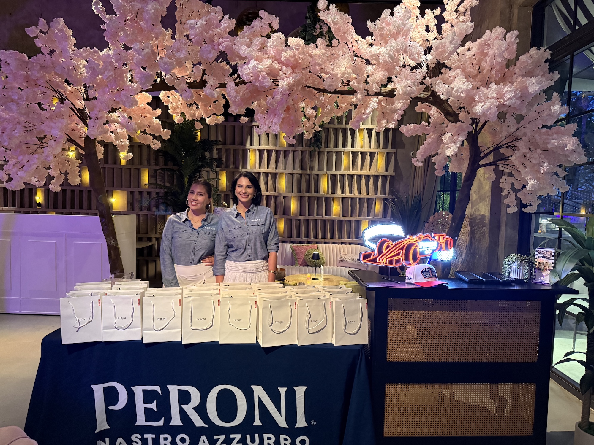 Photo of two women posing inside a bar in front of a table with white goodie bags and a tablecloth reading, "Peroni." A decorative cherry-blossom tree is seen above and behind them