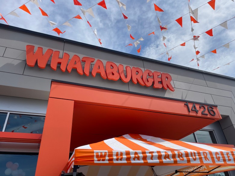 Exterior of a Whataburger zoomed into the name on the building with orange and white decorative flags above the building and a tent below it. The building number (1426) is visible
