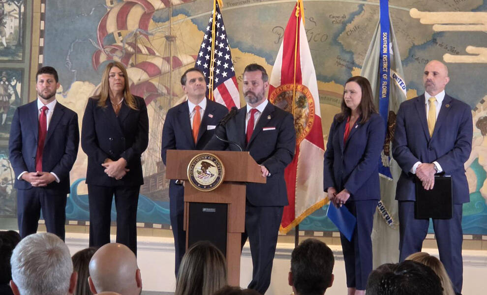 Government officials and law enforcement representatives stand behind a podium during a press conference with U.S. and Florida state flags displayed in the background.