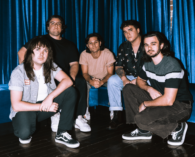 A direct-flash promotional photograph of the five members of the band Rohna posing together indoors. They are arranged in a staggered, tight grouping—with two members crouching in the front, two seated in the middle, and one standing in the back—against a backdrop of bright, shiny blue curtains. The band members are looking directly at the camera and are dressed in casual indie-rock attire, including a grey vest over a white t-shirt, a solid black t-shirt, a light pink short-sleeve shirt, a dark patterned button-down, and a striped t-shirt. The bright flash lighting casts crisp shadows on the blue curtains behind them.