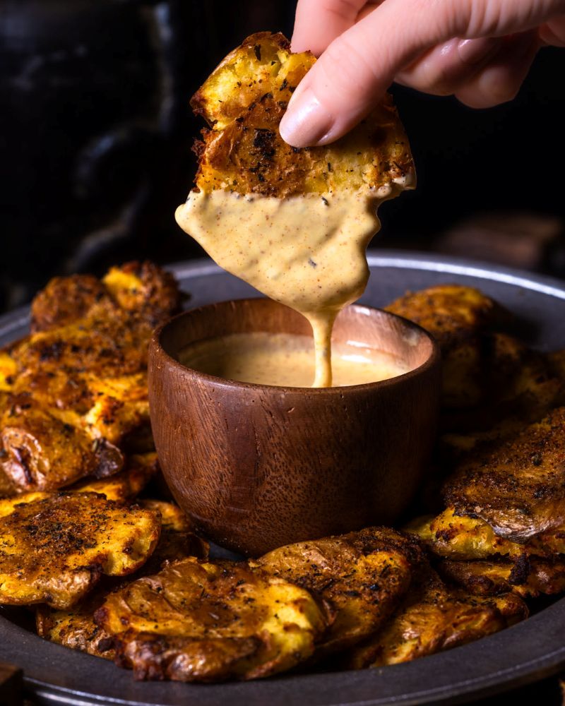 A hand dipping a crispy smashed potato into a wooden bowl filled with the Six Ravens Disney Springs honey mustard sauce, surrounded by a plate of potatoes.