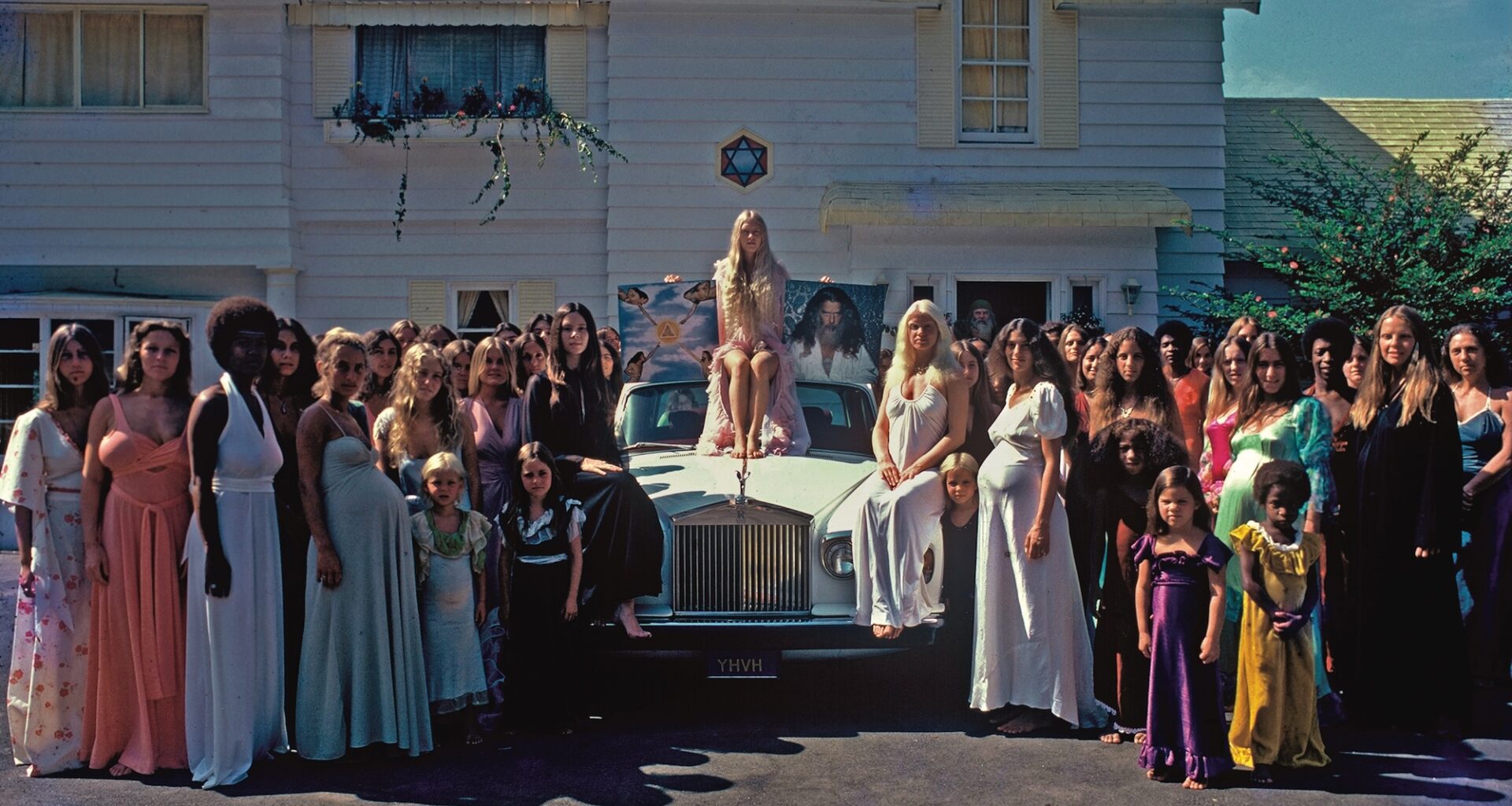 photo depicting a crowd of women around a Rolls-Royce. A woman sits on the car holding a poster depicting a cult leader