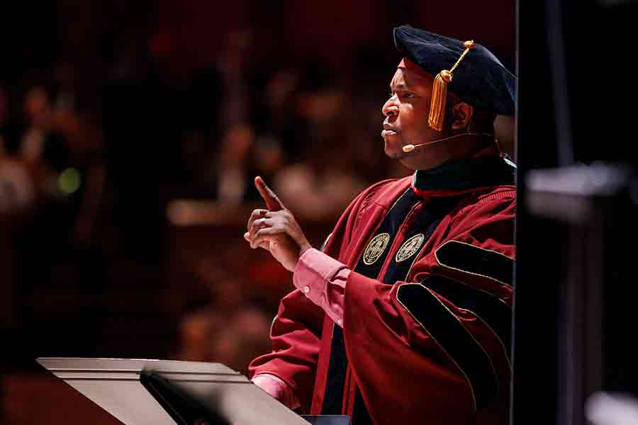 A man in traditional academic cap and gown delivers a speech at a commencement ceremony.