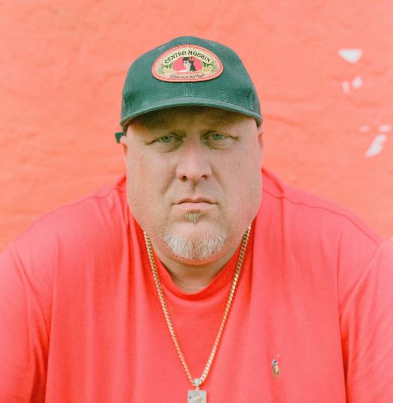 A close-up portrait of a light-skinned person with a goatee standing against a bright, textured coral-red wall. They are wearing a bright red t-shirt that closely matches the background, a gold chain with a square pendant, and a dark green baseball cap. The cap features an oval patch with a vintage graphic of a woman's face and text. The person is looking directly at the camera with a calm, serious expression. The overall lighting is bright and even, highlighting the vibrant color coordination of the outfit and the background.