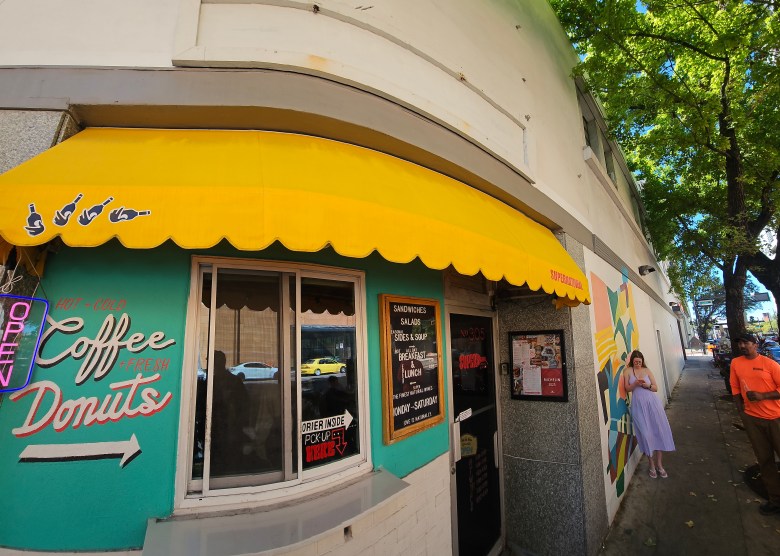 A wide-angle, daytime exterior photograph of the Supernatural Food & Wine storefront in Tampa. The building features a bright yellow scalloped awning extending over a section of wall painted vibrant teal, which displays the words 'Coffee Donuts' in large, red stylized script above a pointing arrow. Beneath the awning is a glass window with a sign reading 'ORDER INSIDE PICK-UP HERE', situated next to a framed wooden menu board and the glass entrance door. To the right of the entrance, the building's white wall is decorated with a colorful geometric mural. The sidewalk is deeply shaded by a large, leafy green tree. Two individuals are visible walking on the sidewalk to the right: one wearing a light purple dress and the other wearing a bright orange shirt and a dark hat. The reflection of street-parked cars is visible in the shop's front window.