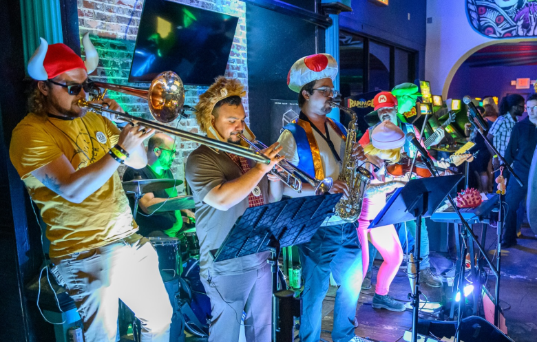 A vibrant, eye-level live concert photograph of the band The Button Smashers performing on stage in front of a brick wall. The band members are dressed in Nintendo-inspired costumes. From left to right: the first member wears a red-horned Viking helmet and sunglasses, playing a trombone. The second member has a fuzzy, lion-like mane hood and is playing a trumpet. The third member wears a red-capped Toad mushroom hat and a blue vest, playing a saxophone. In the background, other band members are visible playing a violin, electric guitar, and drums, illuminated by bright blue stage lighting. The audience is visible on the far right, also partially bathed in cool-toned light.
