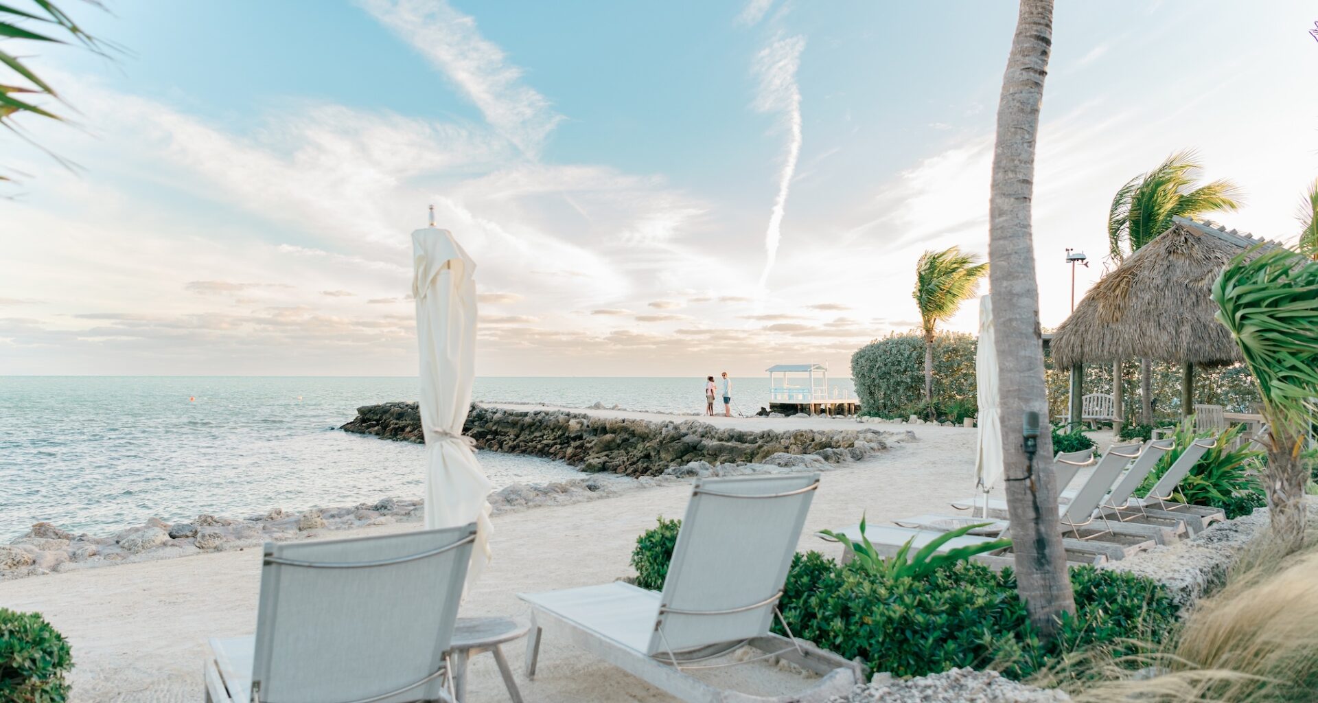 photo of a beach showing two beach chairs and folded beach umbrellas next to a tiki hut with two small people on a jetty in the distance