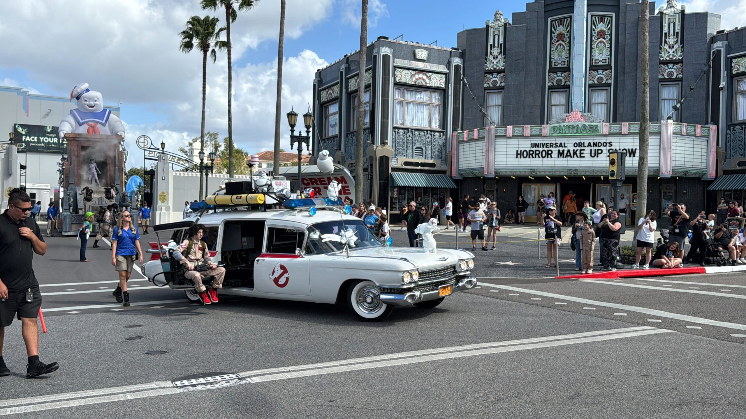 A white car with Ghostbusters logo drives in a parade. People watch from the sidewalk. A Stay Puft Marshmallow Man figure is visible in the background.