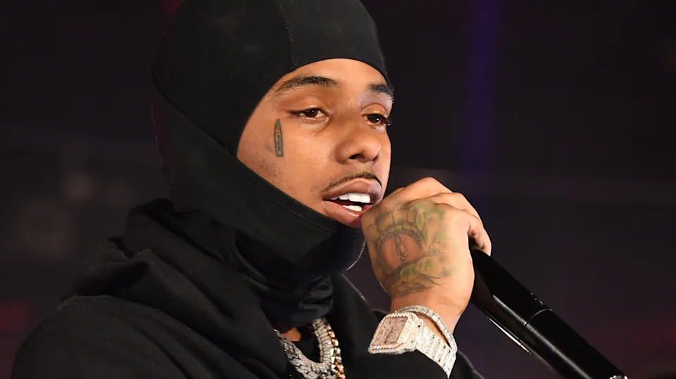 Pooh Shiesty wearing a black hood speaks into a microphone. They have facial tattoos and are wearing a watch and chain.