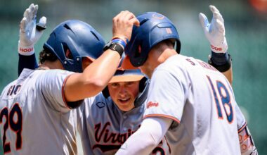 Virginia Cavaliers baseball players in white uniforms celebrate in a huddle on the field, high-fiving each other around a central player.