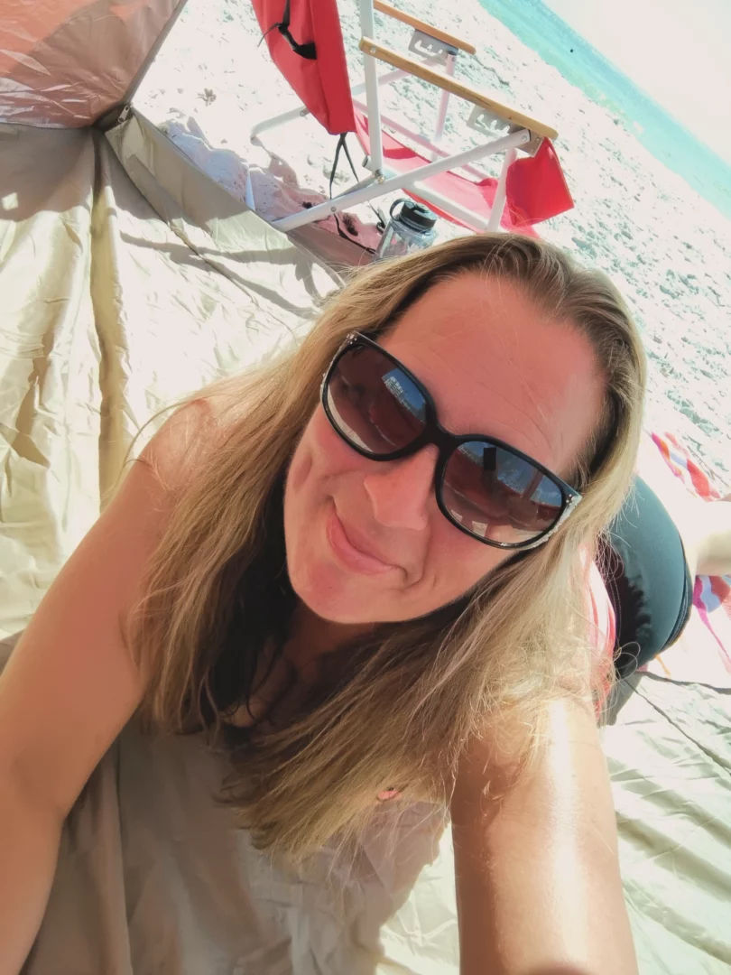 Woman with long hair wearing sunglasses, smiling, at beach tent with ocean and sky in background.